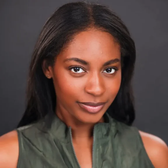 A woman with long dark hair and brown eyes, wearing a sleeveless, high-collar green top, poses in front of a plain dark background.