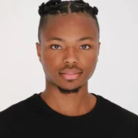 A person with neatly styled short hair in twists, a trimmed goatee, and a black shirt, facing the camera against a plain white background.