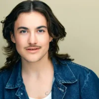 A person with long dark hair, a mustache, and light facial hair wears a denim jacket over a white shirt, looking directly at the camera against a plain beige background.