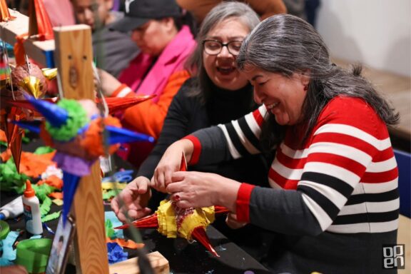 Two women sit at a table crafting colorful piñatas with paper, glue, and decorations, surrounded by materials and other people working.