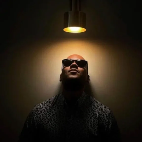 A man wearing sunglasses looks up at a lit ceiling lamp, casting a circular glow above his head in an otherwise dark room.