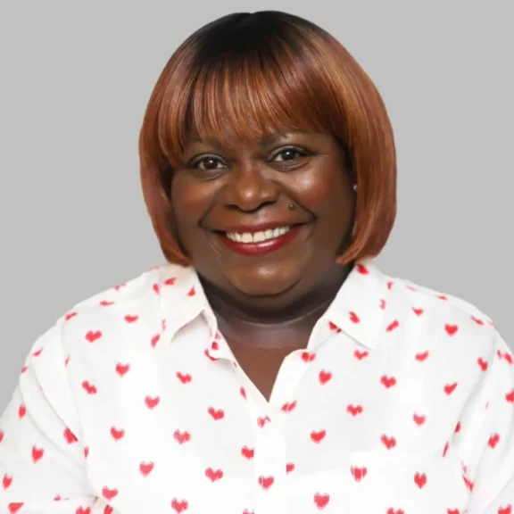 A woman with a short, straight, reddish-brown hairstyle smiles, wearing a white blouse with red heart patterns, in front of a plain grey background.