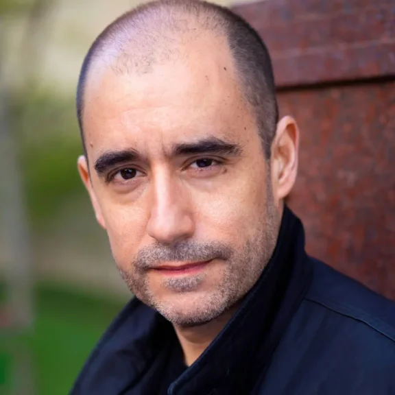 A man with a shaved head and slight stubble looks into the camera, wearing a black jacket, standing in front of a blurred outdoor background.