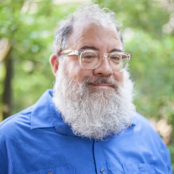 A man with gray hair, a full gray beard, and glasses is wearing a blue shirt and standing outdoors with greenery in the background.
