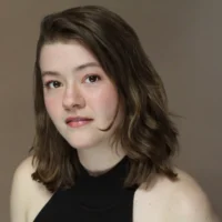 A woman with shoulder-length brown hair and fair skin looks at the camera, wearing a sleeveless black top against a plain brown background.