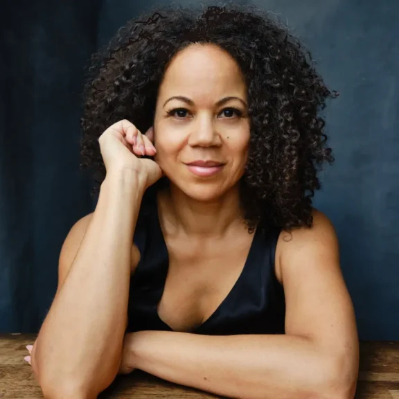 A woman with curly hair sits at a wooden table, resting her chin on her hand and looking at the camera, against a dark background.