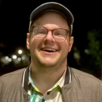 A man with glasses and a cap smiles at the camera at night. He is wearing a brown jacket over a patterned shirt. The background is dark with blurred lights.