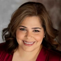Woman with wavy brown hair wearing a red top, smiling at the camera with a neutral background.