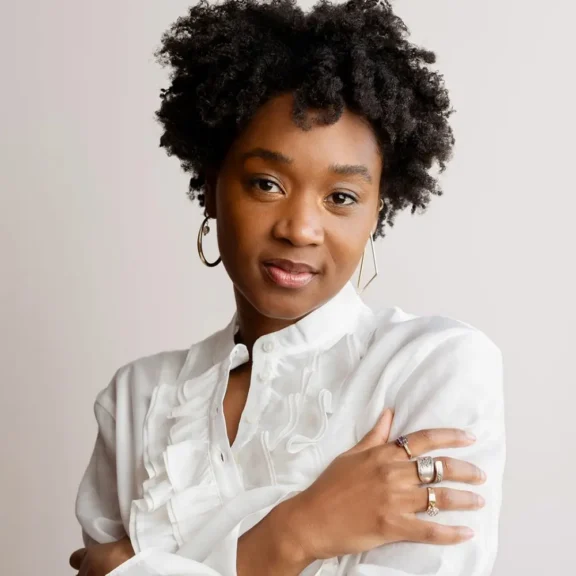 A woman with short curly hair, wearing a white ruffled blouse and hoop earrings, poses with her arms crossed and a neutral expression against a plain background.