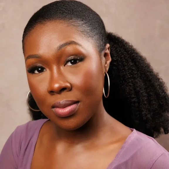A woman with dark, curly hair pulled back into a ponytail, wearing hoop earrings and a lavender top, looks at the camera against a neutral background.
