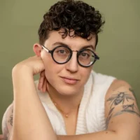 Person with short curly hair, round glasses, and visible tattoos on their arm, wearing a white sleeveless top, sits with their chin resting on one hand against a plain background.