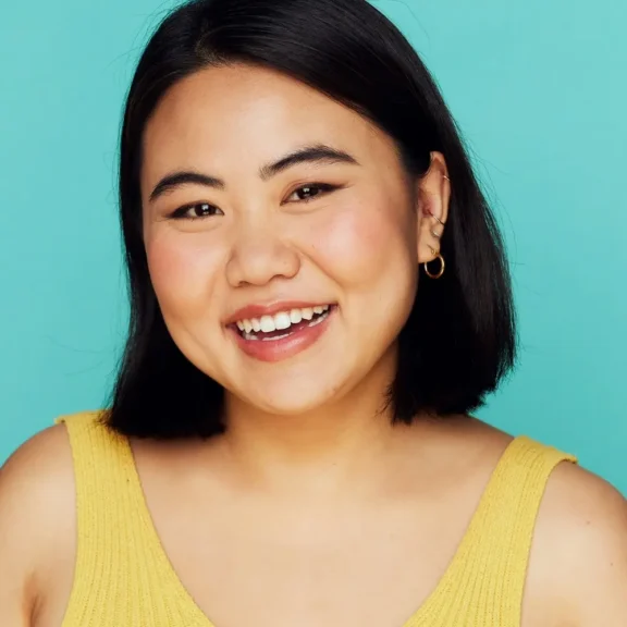 A person with short black hair wearing a yellow sleeveless top smiles in front of a turquoise background.