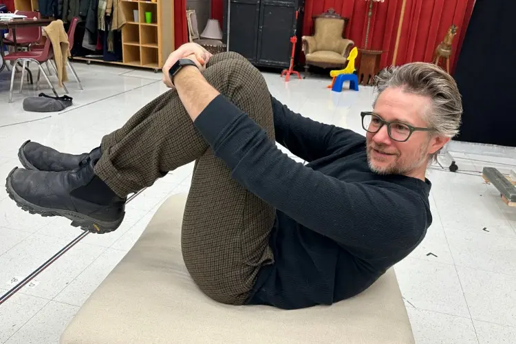 A man with glasses and a beard sits on a beige bench, hugging his knees to his chest, in a room with red curtains, chairs, and various items in the background.