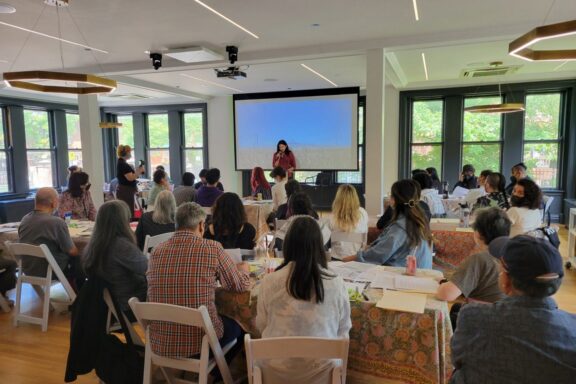 A group of people sit at tables in a bright room, watching a presenter speaking in front of a large screen displaying an outdoor scene.