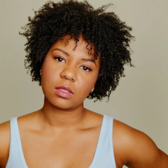 A woman with short curly hair wearing a light blue tank top looks directly at the camera with a neutral expression.