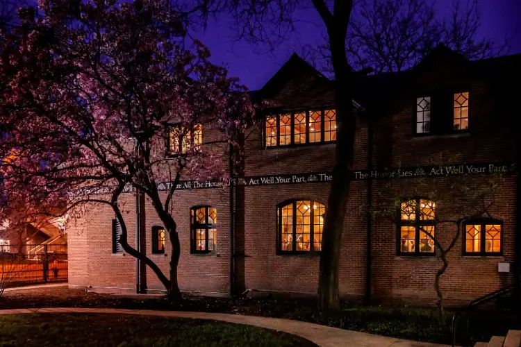 A brick building with lit windows at night, surrounded by blooming trees and a curved pathway under a deep blue sky.