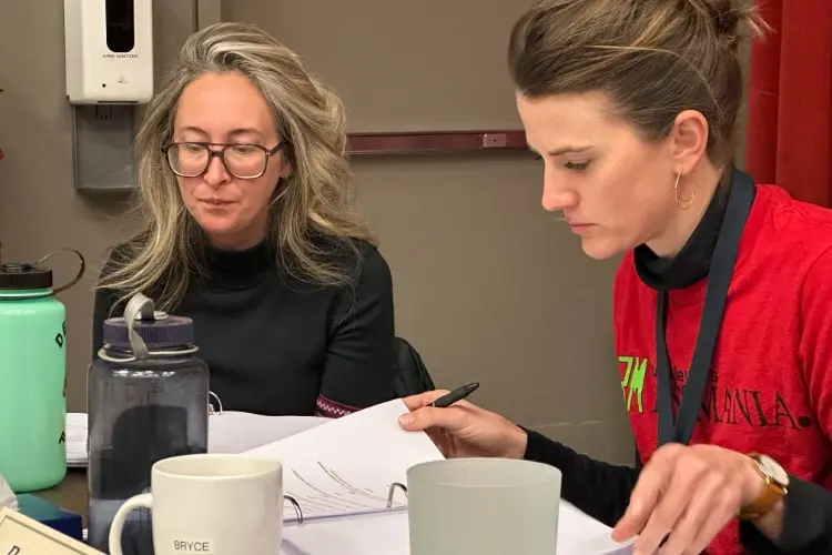 Two women sit at a table reviewing documents, with several mugs and water bottles in front of them.