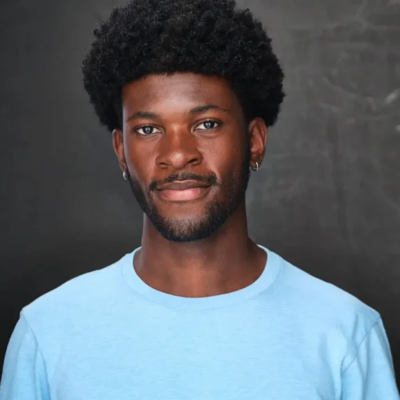 A man with short curly hair, a beard, and hoop earrings wears a light blue shirt and stands in front of a dark, plain background.