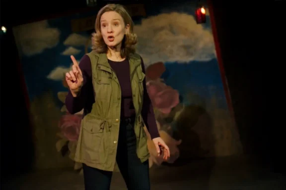 A woman stands on stage, holding up one finger while speaking, with a colorful painted backdrop behind her.