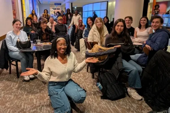 A group of people, mostly young adults, sit and stand together in a restaurant. One person kneels at the front, smiling with arms open. Everyone appears to be enjoying themselves.