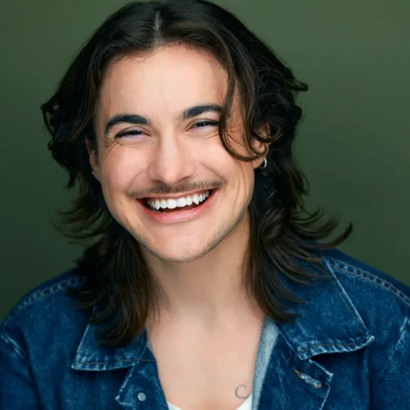 Person with long brown hair and a mustache smiling broadly, wearing a denim jacket over a light shirt, in front of a plain green background.