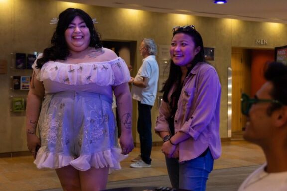 Two people stand and smile indoors near a seated person; one wears a white off-shoulder dress, the other a tan shirt. A man walks past in the background near a "MEN" restroom sign.