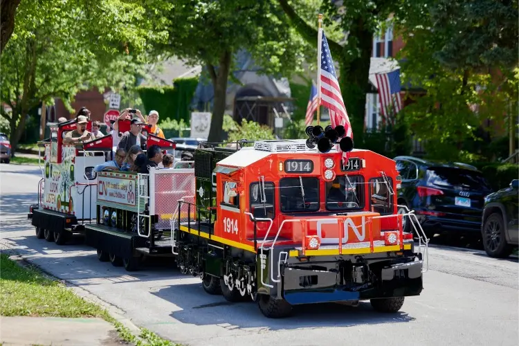 A small train decorated with an American flag carries passengers along a residential street lined with trees and parked cars.