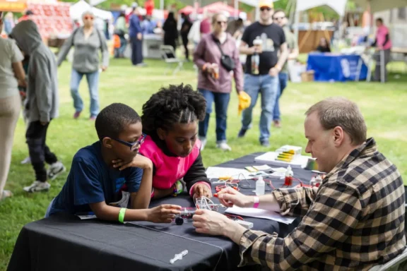 An adult demonstrates a science experiment to two children at an outdoor event, with people and booths visible in the background.