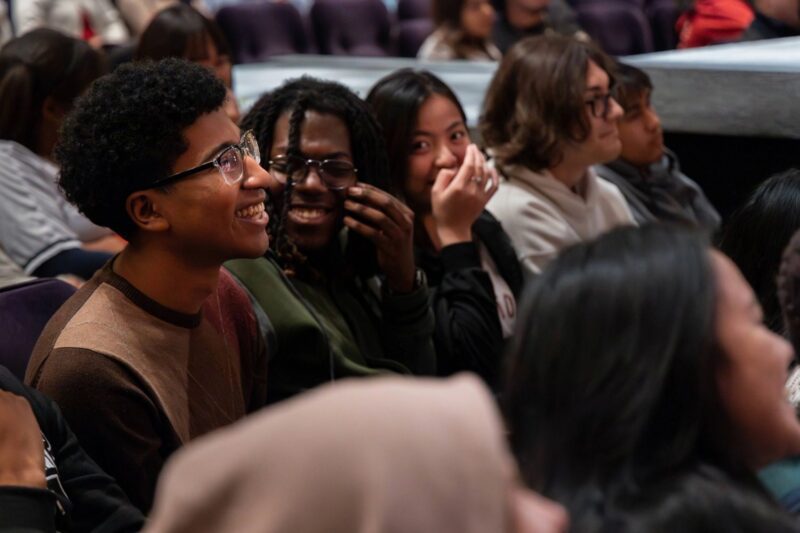 A group of young people sit closely together in an audience, smiling and laughing while watching an event or performance.