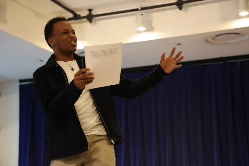 A person holds a sheet of paper and gestures expressively while speaking in a room with dark blue curtains and spotlights overhead.