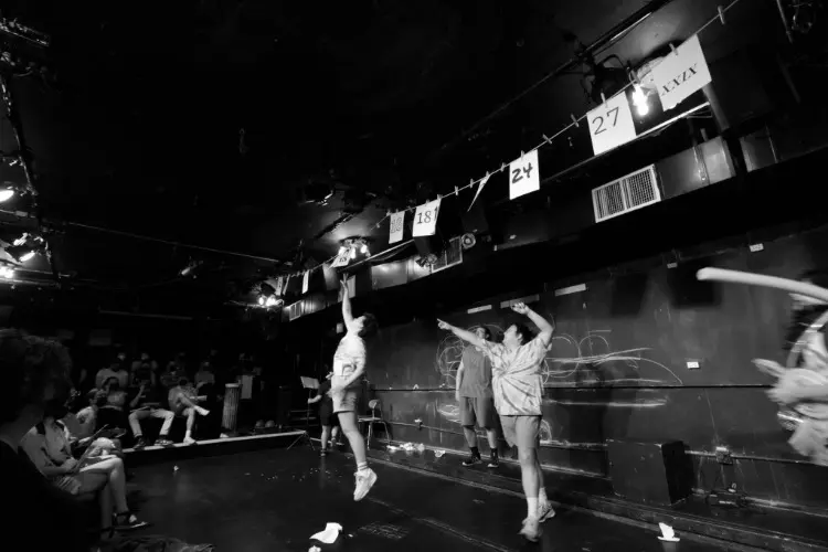 A group of performers in costume jump and reach toward hanging numbered papers on a string in a dimly lit theater as an audience watches.