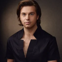 A man with medium-length brown hair and blue eyes wearing a black shirt with a unique square neckline poses against a neutral background.