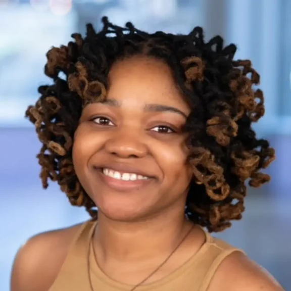 A woman with short, curly two-tone hair smiles while looking at the camera, wearing a tan sleeveless top and a necklace, with a blurred background.