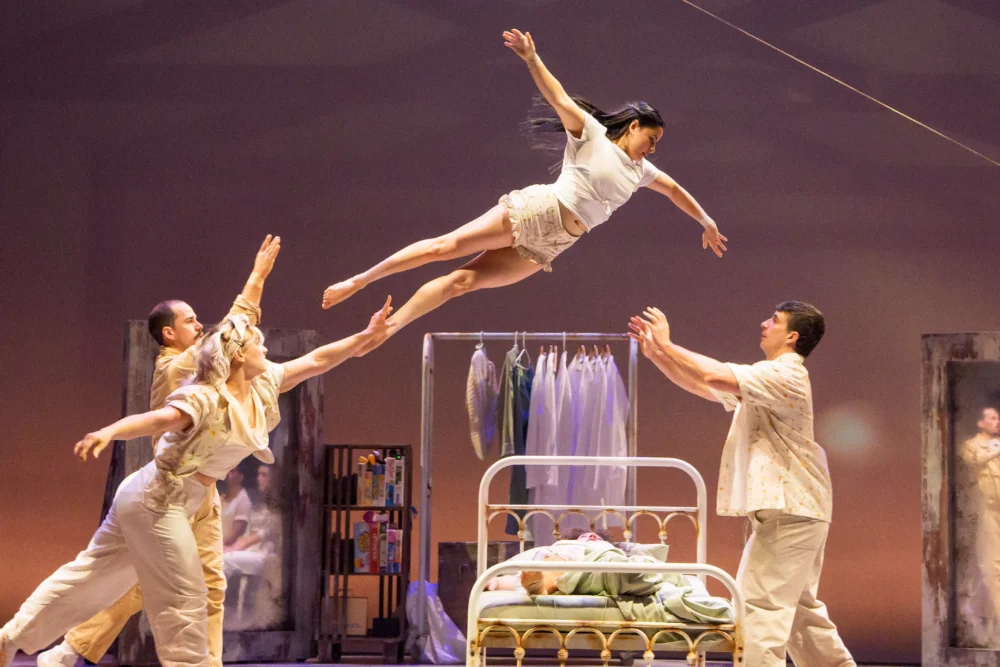 Four performers catch a woman flying horizontally above a bed on stage; costumes and clothes hang in the background.