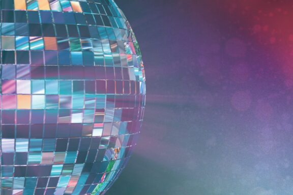 Close-up of a shiny disco ball reflecting colorful lights against a blurred, purple and red background.