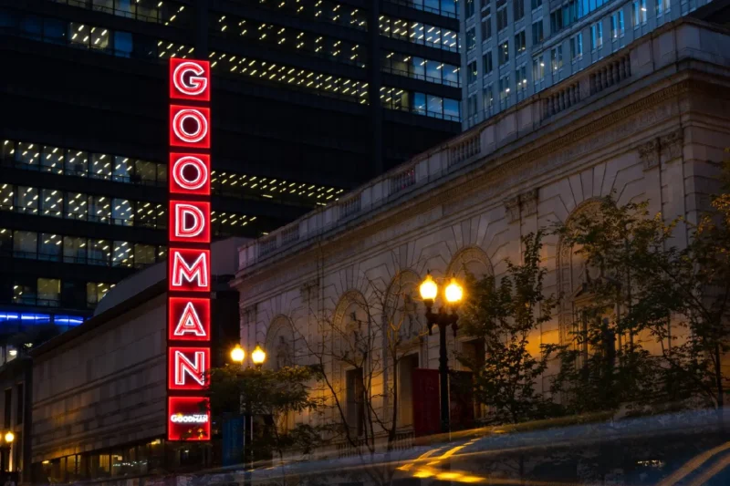 A red neon "GOODMAN" vertical sign is lit on the exterior of a theater building at dusk, with modern office buildings in the background.