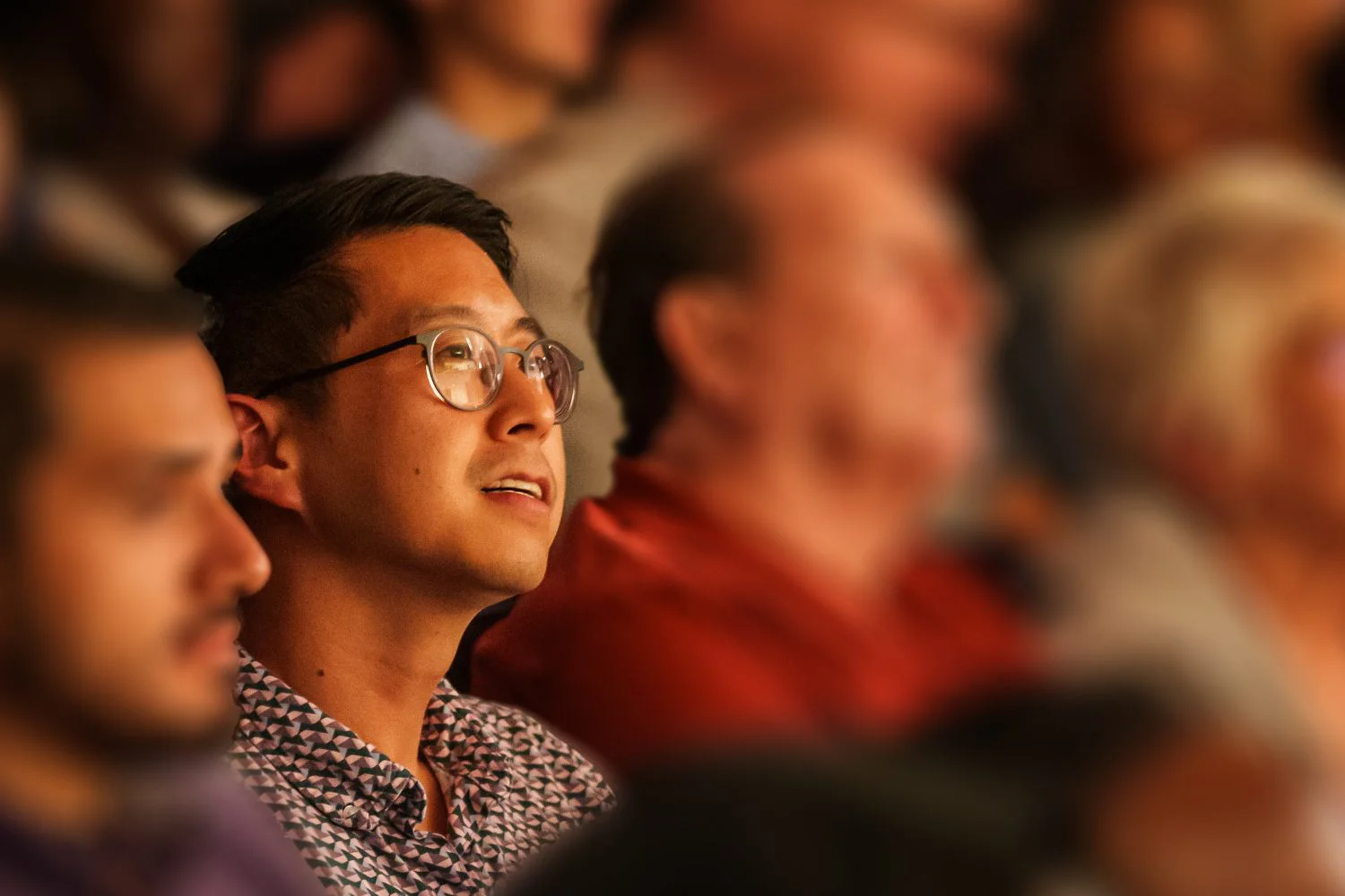 A man wearing glasses and a patterned shirt sits in an audience, attentively watching something off-camera. People around him are blurred in the background.