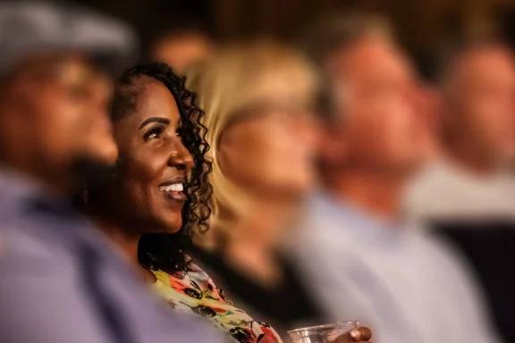 A woman with curly hair smiles while holding a cup, sitting among a group of people who are out of focus.