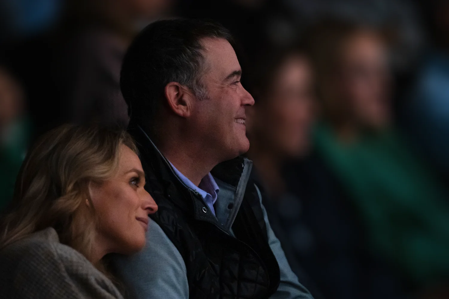 A woman leans her head on a man's shoulder while they both look ahead and smile, surrounded by a blurred crowd in the background.