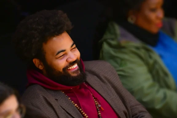 A man with a beard and afro hair, wearing a maroon hoodie and brown blazer, smiles while seated among others in an indoor setting.