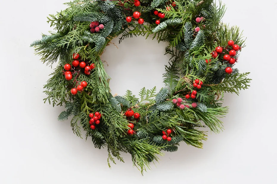 A green wreath made of pine branches decorated with clusters of red berries, hanging against a plain white background.