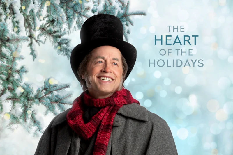 A smiling man in a tall black hat, red scarf, and gray coat stands in front of snowy branches. Text reads: “The Heart of the Holidays.”.