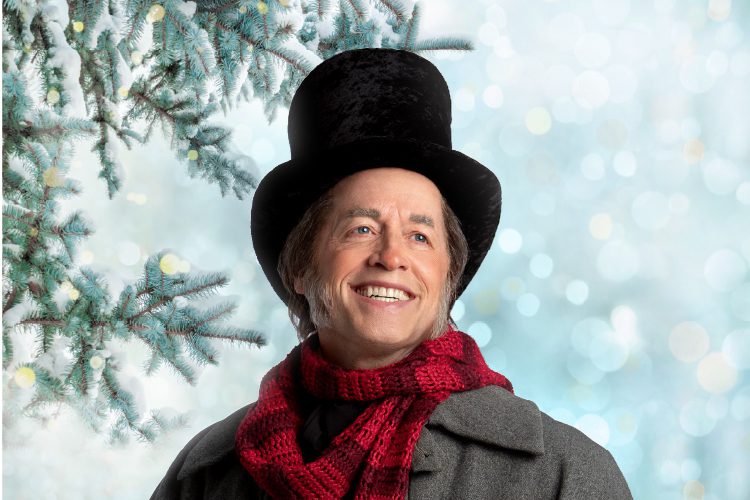 A smiling person in a tall black hat, red scarf, and gray coat stands in front of snow-covered pine branches with a snowy background.