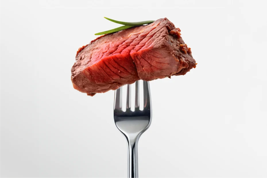 A piece of medium-rare steak with a sprig of rosemary is pierced by a fork against a plain white background.