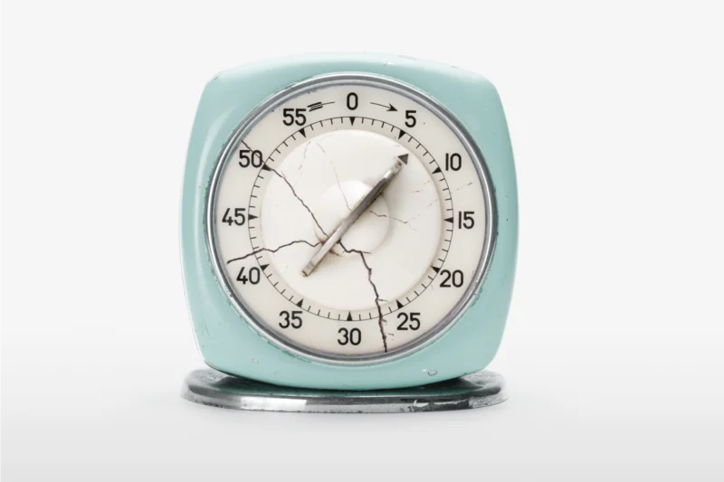 A vintage kitchen timer with cracked glass sits on a white surface, showing approximately 14 minutes.