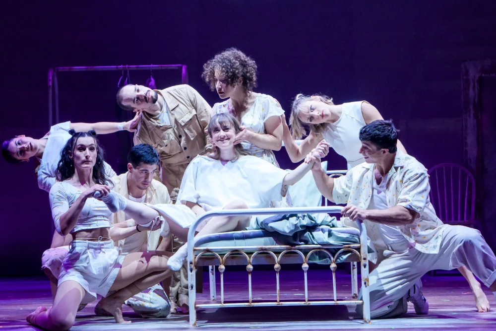 A group of actors in white costumes surround a woman sitting on a hospital bed on stage, set against a purple-lit background.