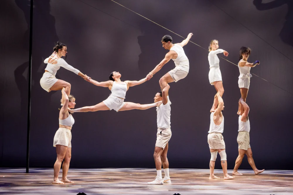 Eight acrobats in white outfits perform a balancing act, forming human towers and holding poses on a stage with a neutral background.