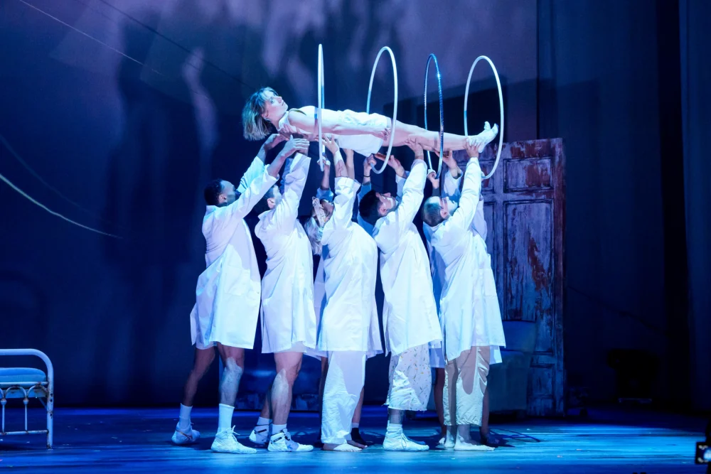 Six people in white outfits hold up rings and support a person in a dress, suspended horizontally above the stage, during a theatrical performance.