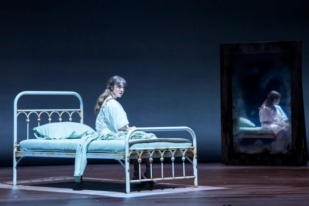 A woman in white sits on a bed, looking toward a large mirror that reflects her image on a dimly lit stage.