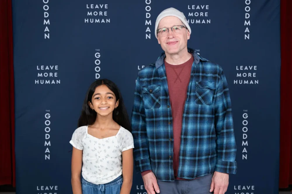 A young girl and an adult man stand together in front of a blue backdrop with repeating text that reads “Leave More Human” and “The Goodman.”